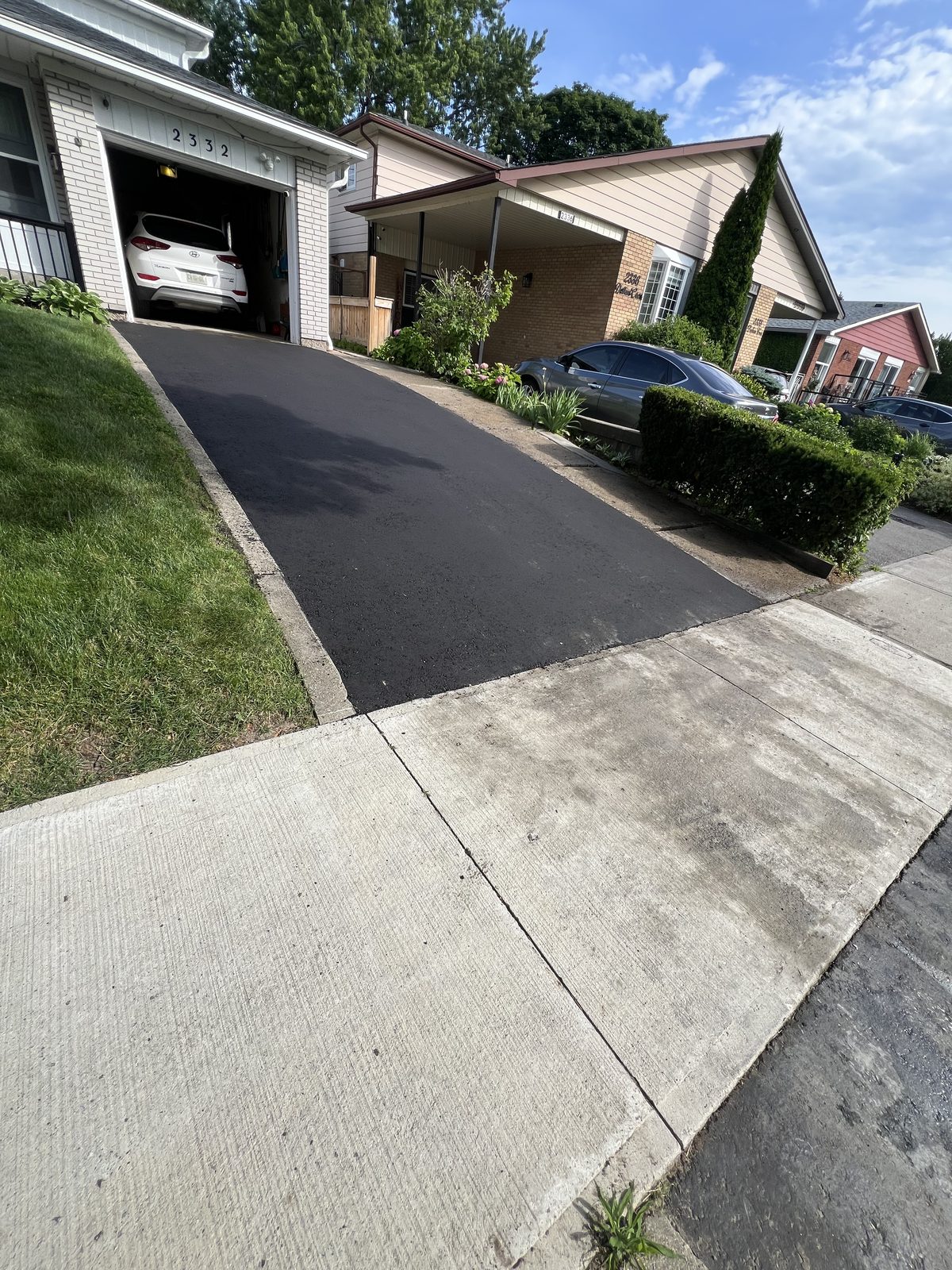 Newly paved smooth black asphalt driveway leading to a garage at a GTA home, bordered by green lawn and concrete walkway
