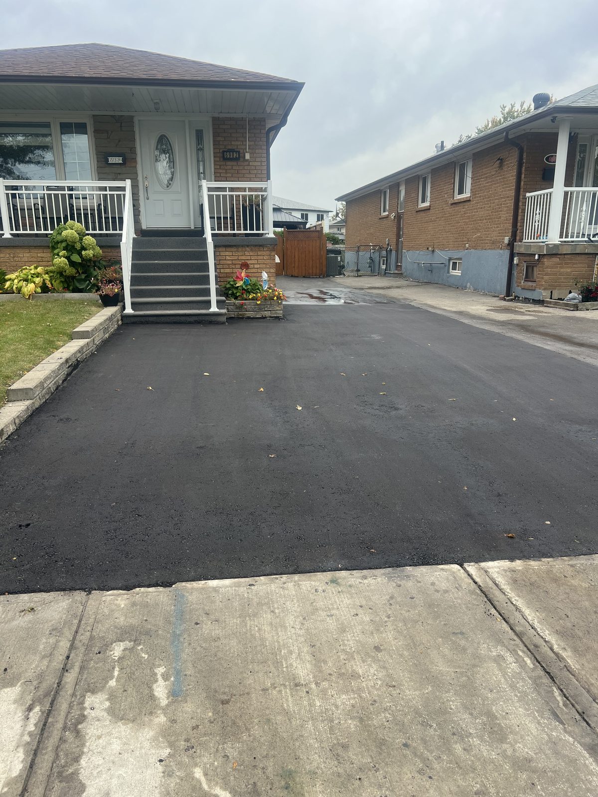 Newly paved smooth black asphalt residential driveway between two brick homes in the GTA, finished at concrete sidewalk