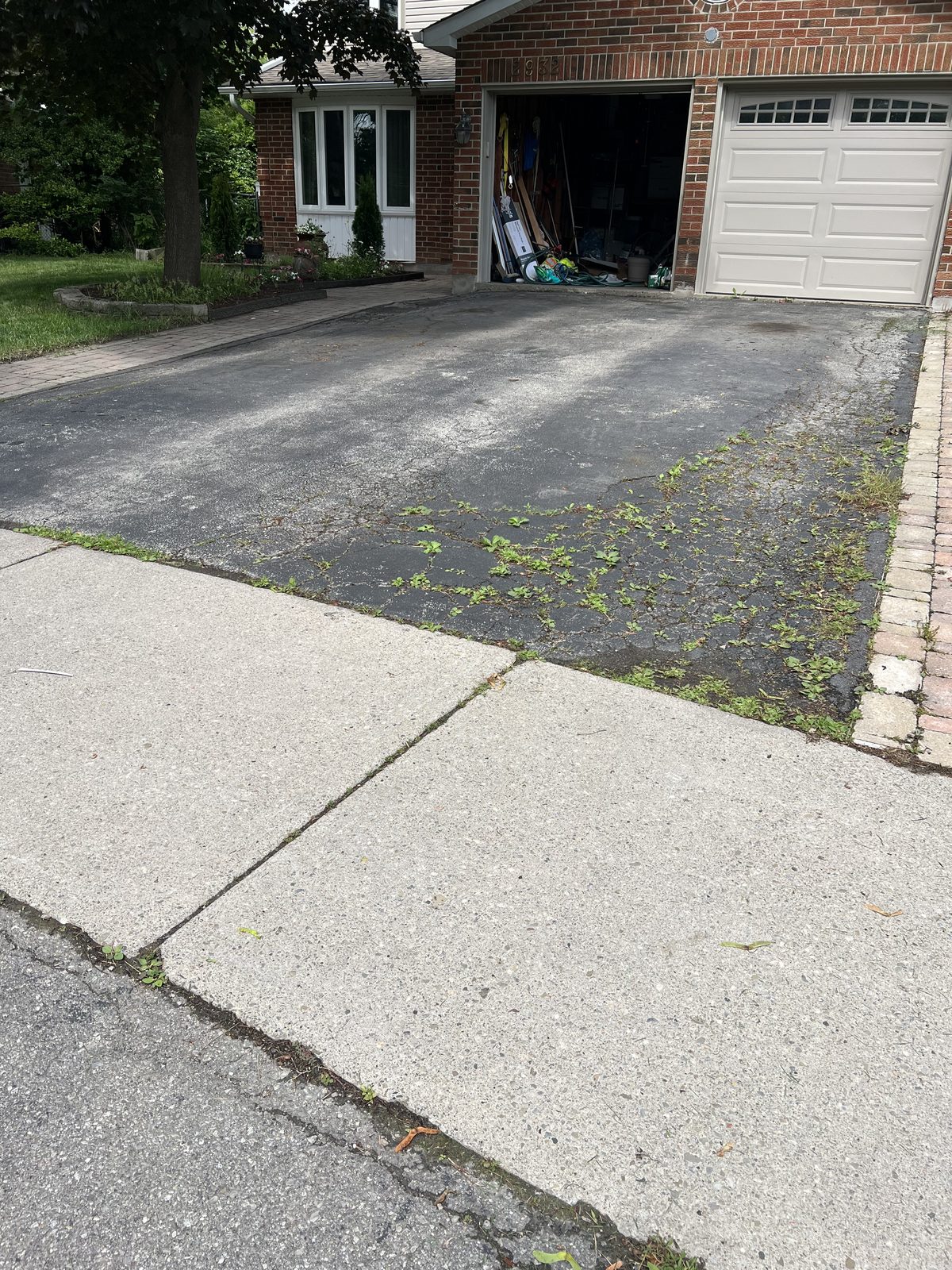 Aging residential asphalt driveway with weeds and surface deterioration beside brick home in the GTA, awaiting repaving