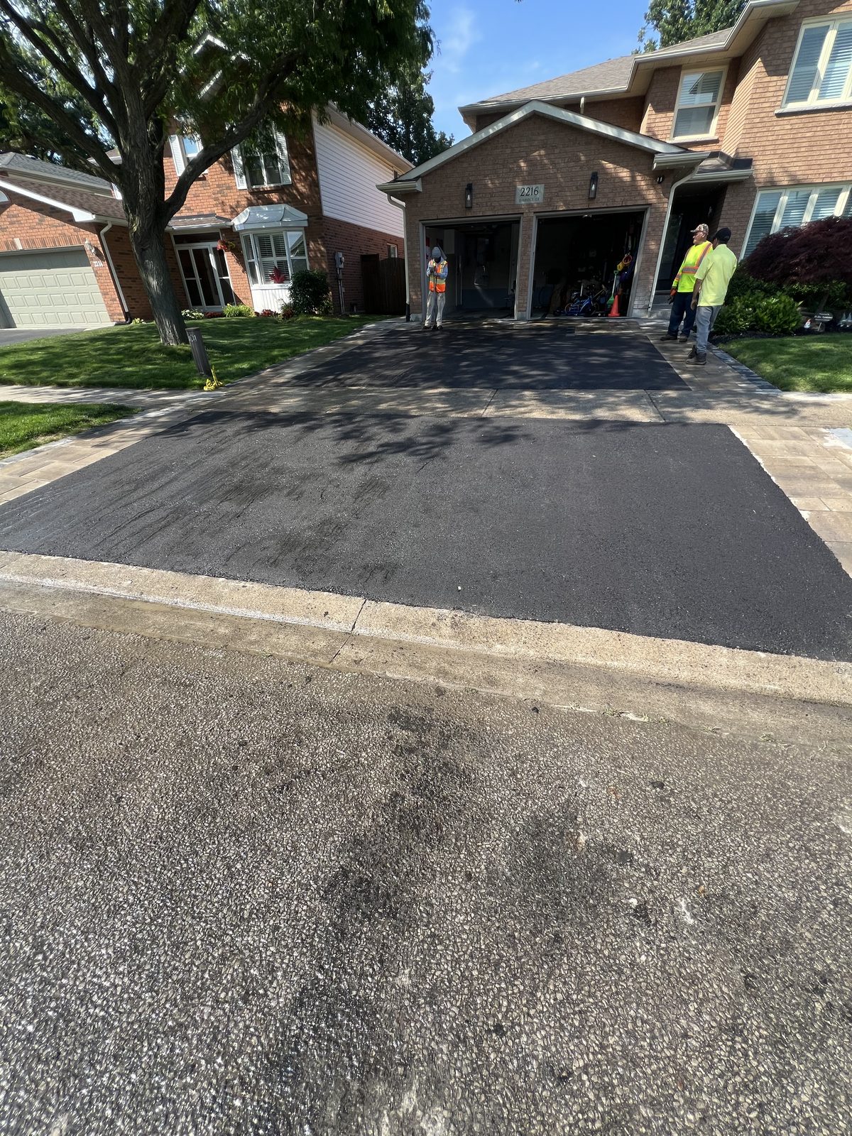 Newly paved asphalt driveway with decorative paver borders at a two-storey brick home in the GTA, crew in safety vests on site